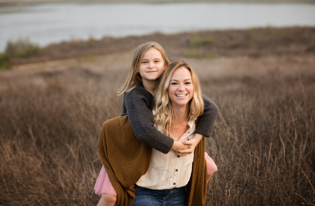 A mom giving her daughter a piggy back ride at a Newport Beach family photo shoot