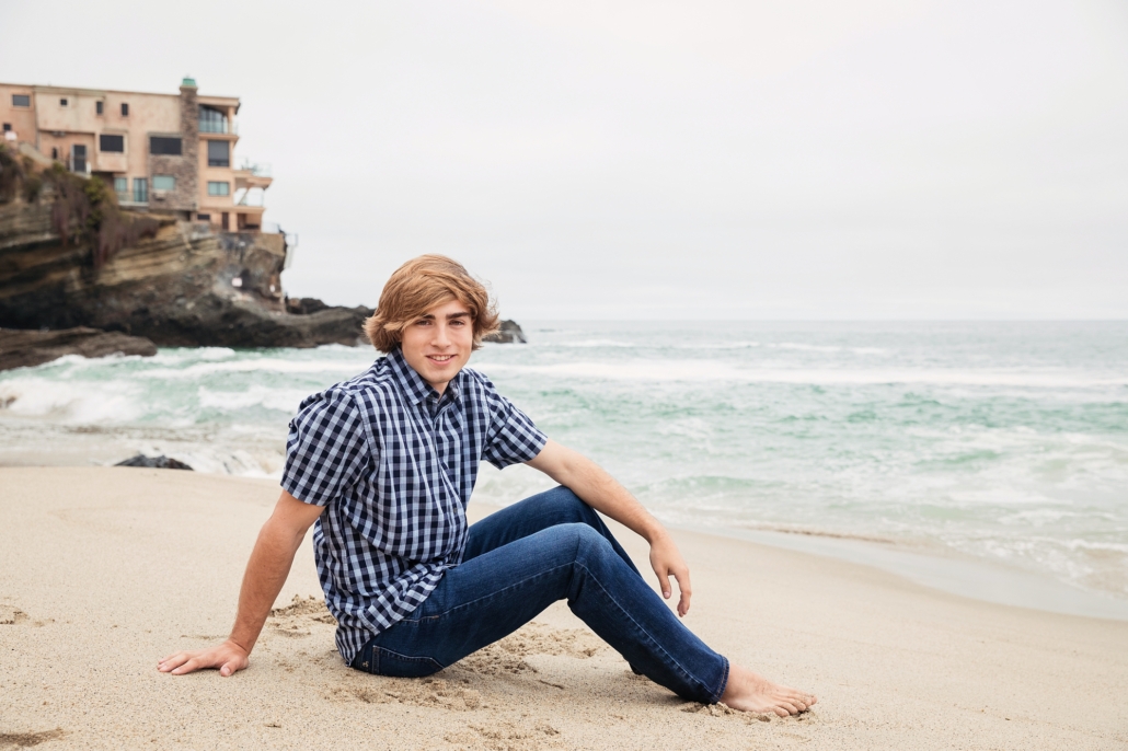 High School senior guy sitting on the beach with an ocean background.