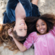 Mother and daughter lying on the beach, heads close together, smiling up at the sky as the sun casts a warm glow on them.