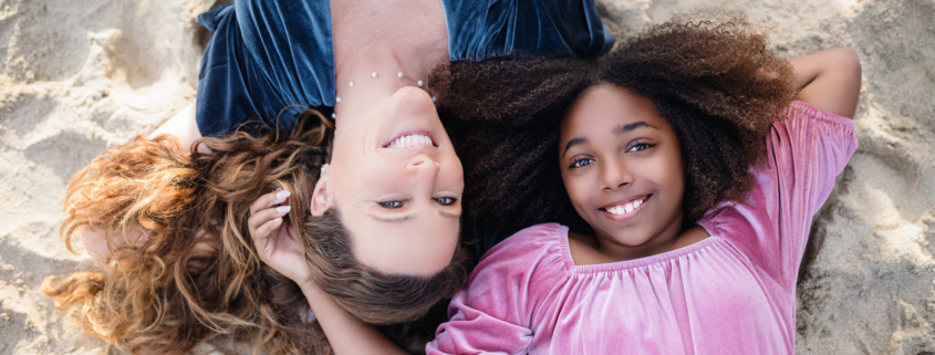 Mother and daughter lying on the beach, heads close together, smiling up at the sky as the sun casts a warm glow on them.