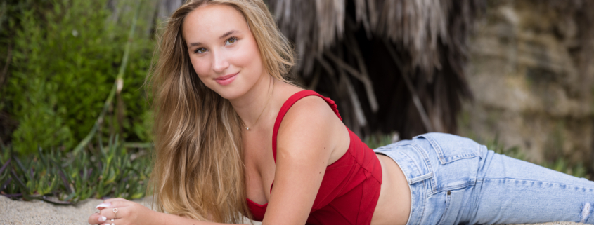 A high school senior girl with long blonde hair poses on the sandy beach, leaning on her elbows and smiling softly. She wears a stylish red crop top and light-wash jeans, with a natural, beachy backdrop of palm fronds and greenery behind her.