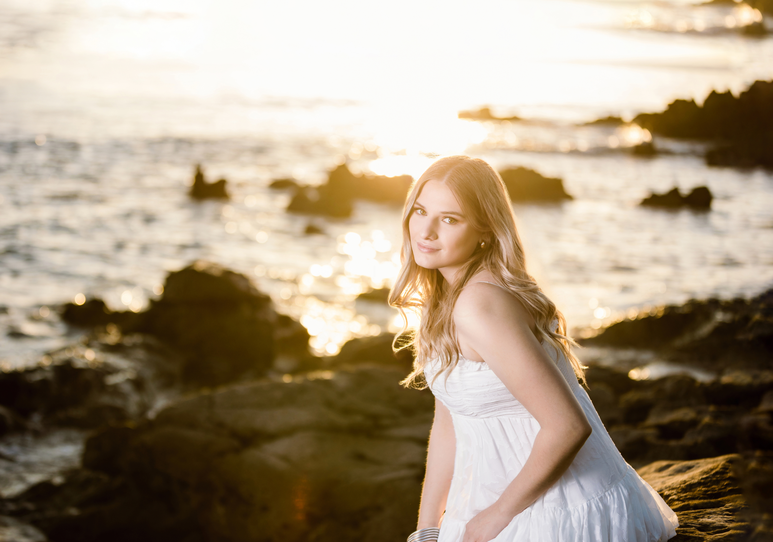 Caldwell sitting on a rocky beach at sunset, glowing in golden hour light wearing a white dress, with soft waves in the background.