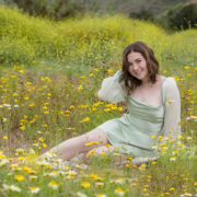 Senior girl sitting in the middle of a blooming wildflower meadow, surrounded by yellow and white daisies, wearing a sage green dress and smiling at the camera.