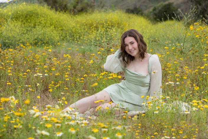 Senior girl sitting in the middle of a blooming wildflower meadow, surrounded by yellow and white daisies, wearing a sage green dress and smiling at the camera.