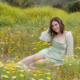 Senior girl sitting in the middle of a blooming wildflower meadow, surrounded by yellow and white daisies, wearing a sage green dress and smiling at the camera.