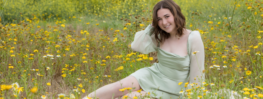 Senior girl sitting in the middle of a blooming wildflower meadow, surrounded by yellow and white daisies, wearing a sage green dress and smiling at the camera.
