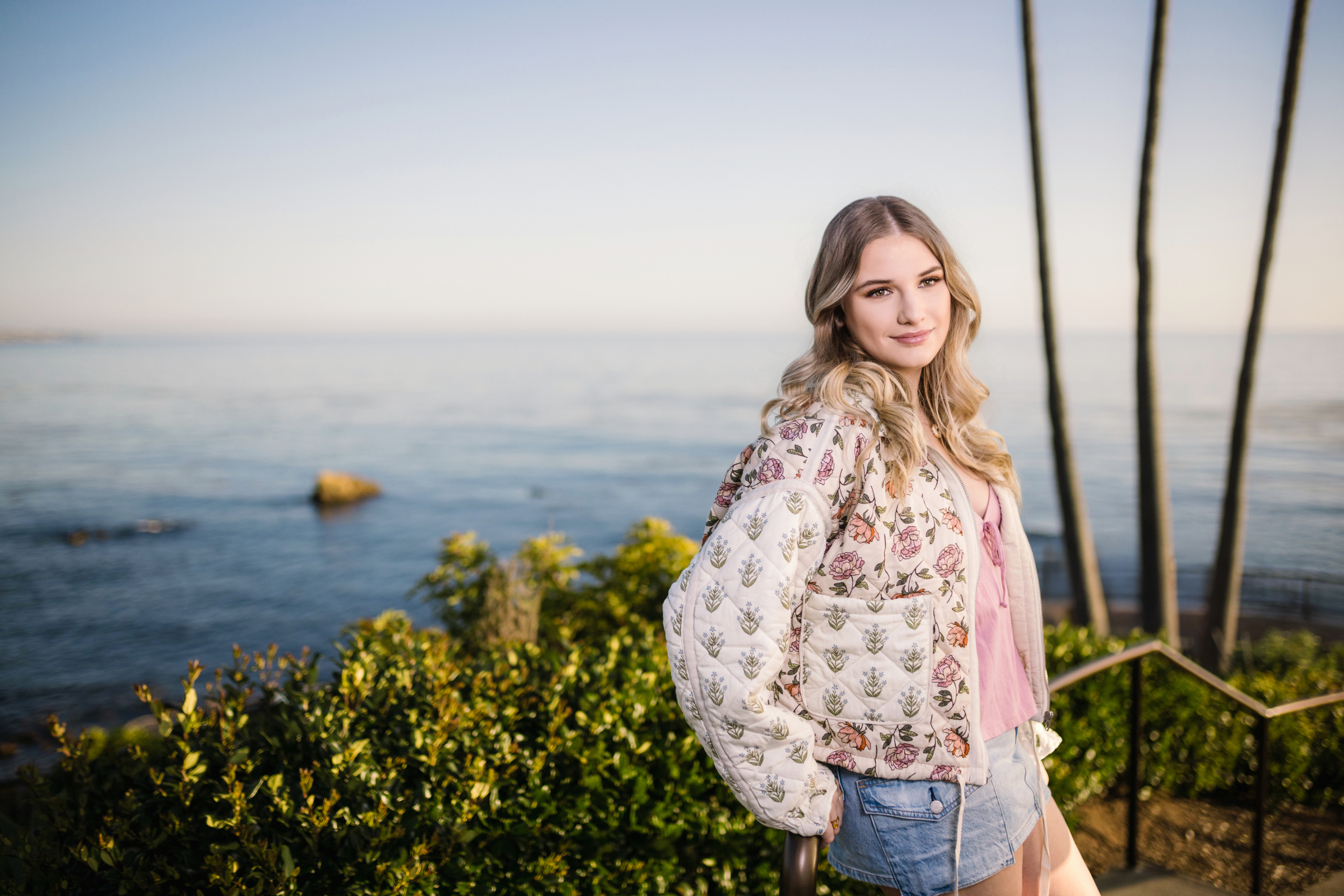 Caldwell smiling in a floral jacket and denim shorts, standing on a staircase overlooking the calm ocean and palm trees in Laguna Beach.