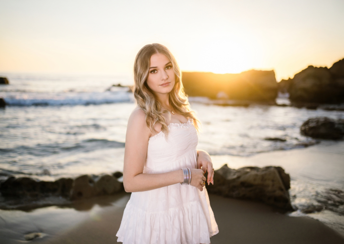 Caldwell standing on the beach in a white dress at golden hour, with soft curls and bracelets catching the warm sunlight.