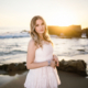 Caldwell standing on the beach in a white dress at golden hour, with soft curls and bracelets catching the warm sunlight.