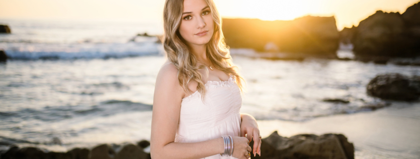 Caldwell standing on the beach in a white dress at golden hour, with soft curls and bracelets catching the warm sunlight.