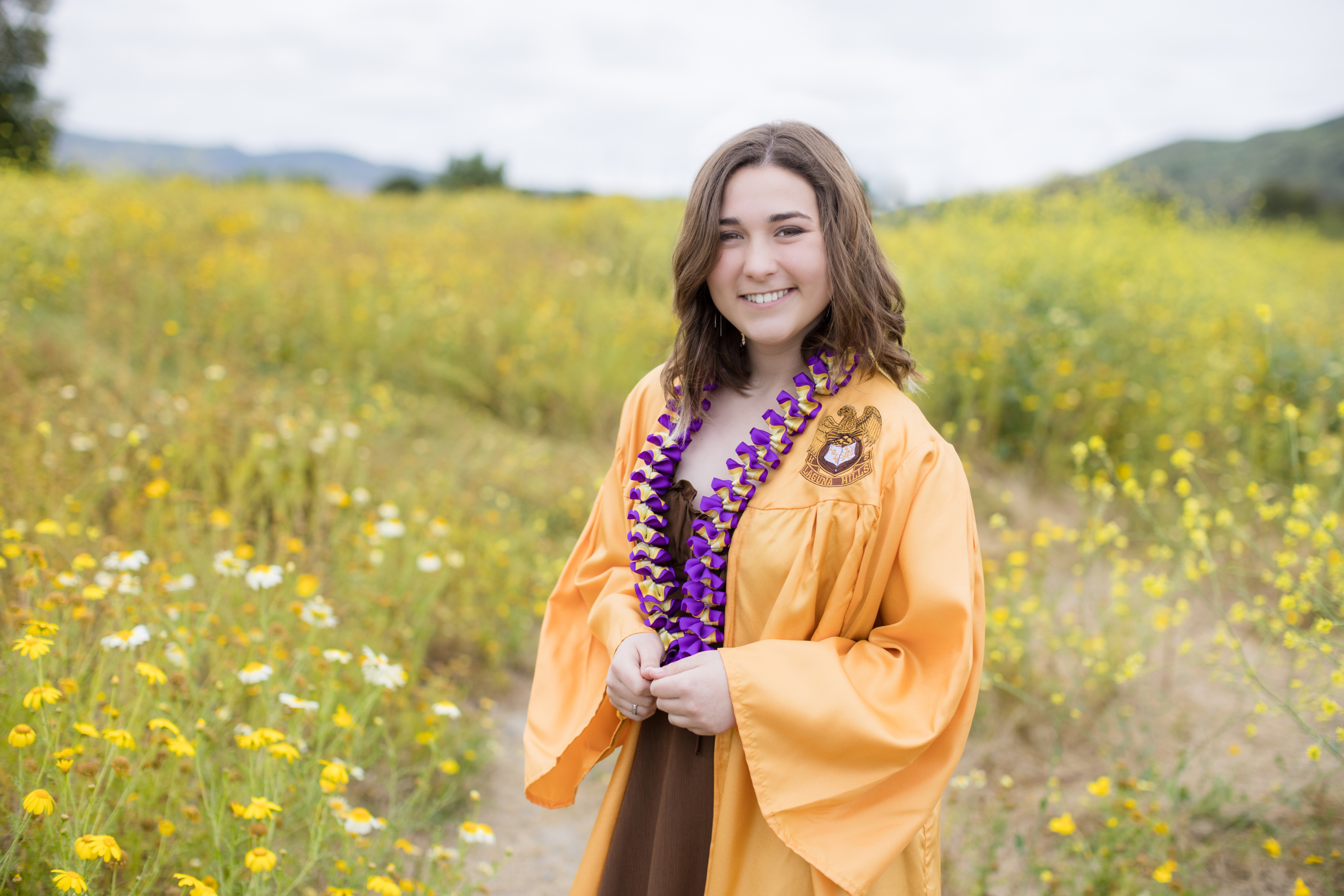 High school senior girl beaming in a field of wildflowers, dressed in her gold graduation gown and purple lei, celebrating her achievement.