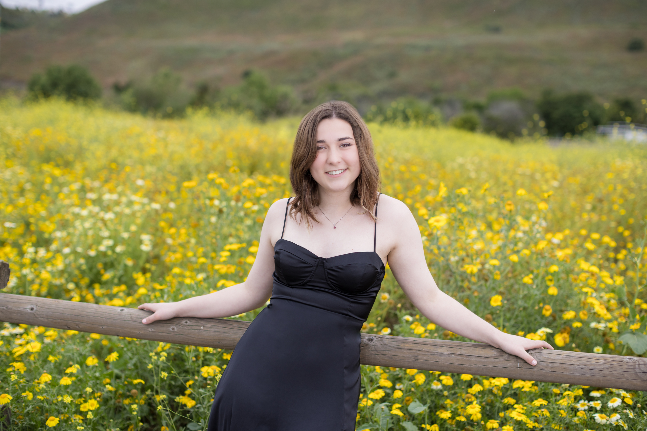 Senior girl in a black dress leaning on a rustic wooden fence with a sea of yellow wildflowers and rolling hills behind her.
