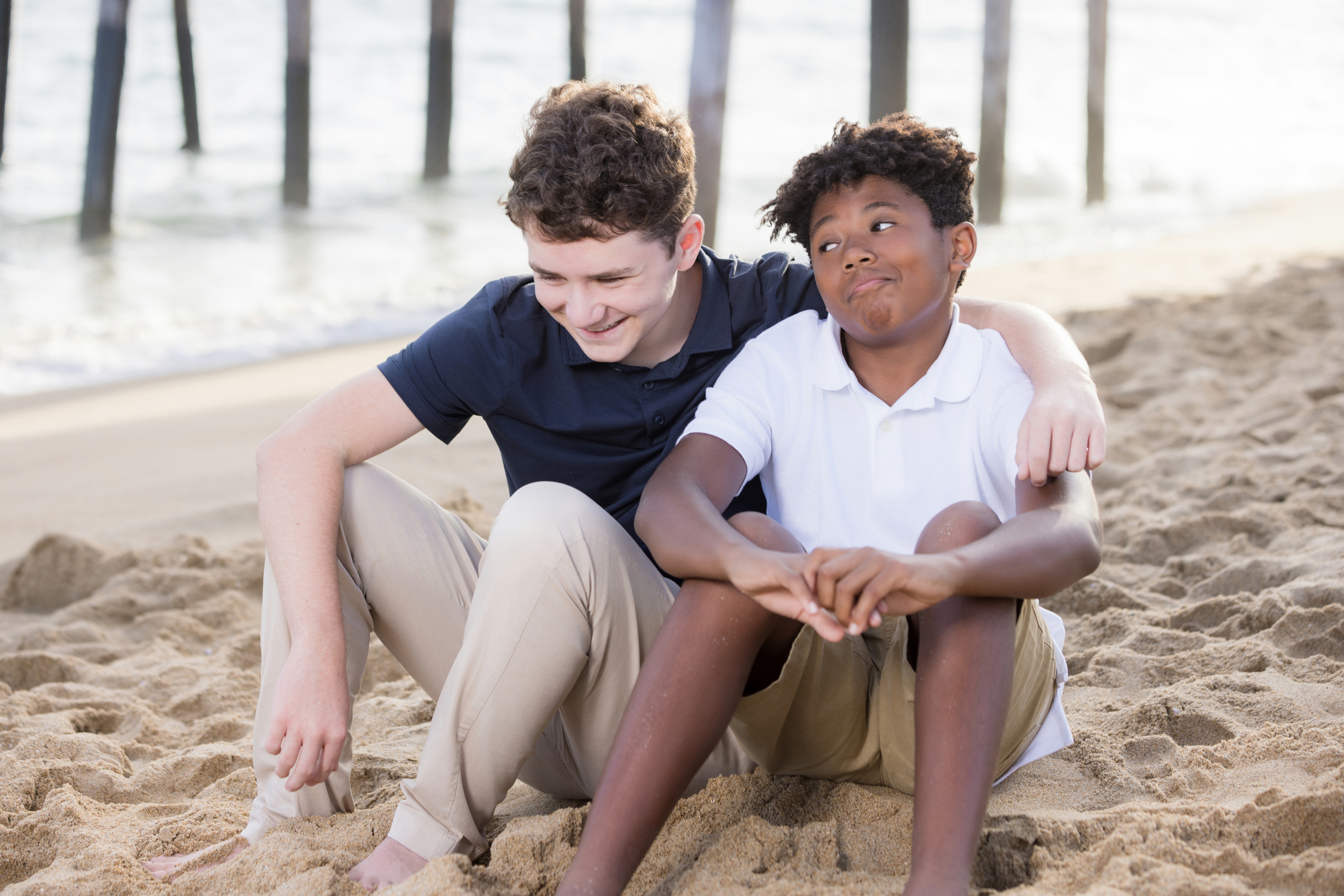 Two boys sitting barefoot on the beach, sharing a lighthearted moment with one making a funny face and the other laughing, with a pier and ocean in the background.