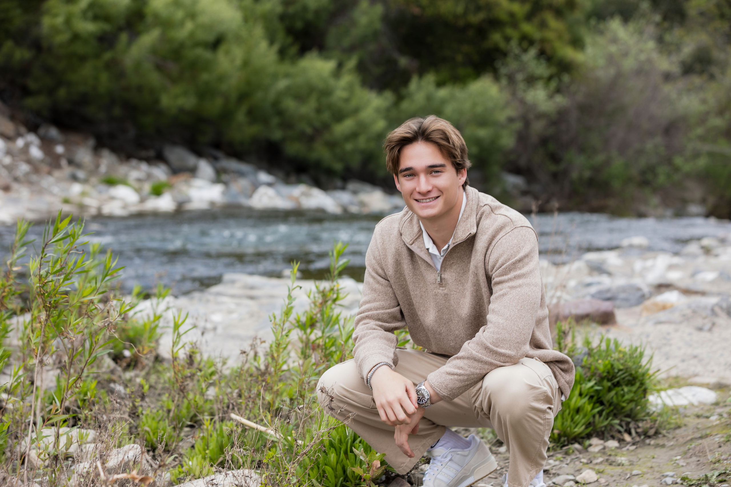 High school senior guy kneeling by a rocky riverbank, dressed in a tan pullover and khaki pants, with soft green foliage in the background.