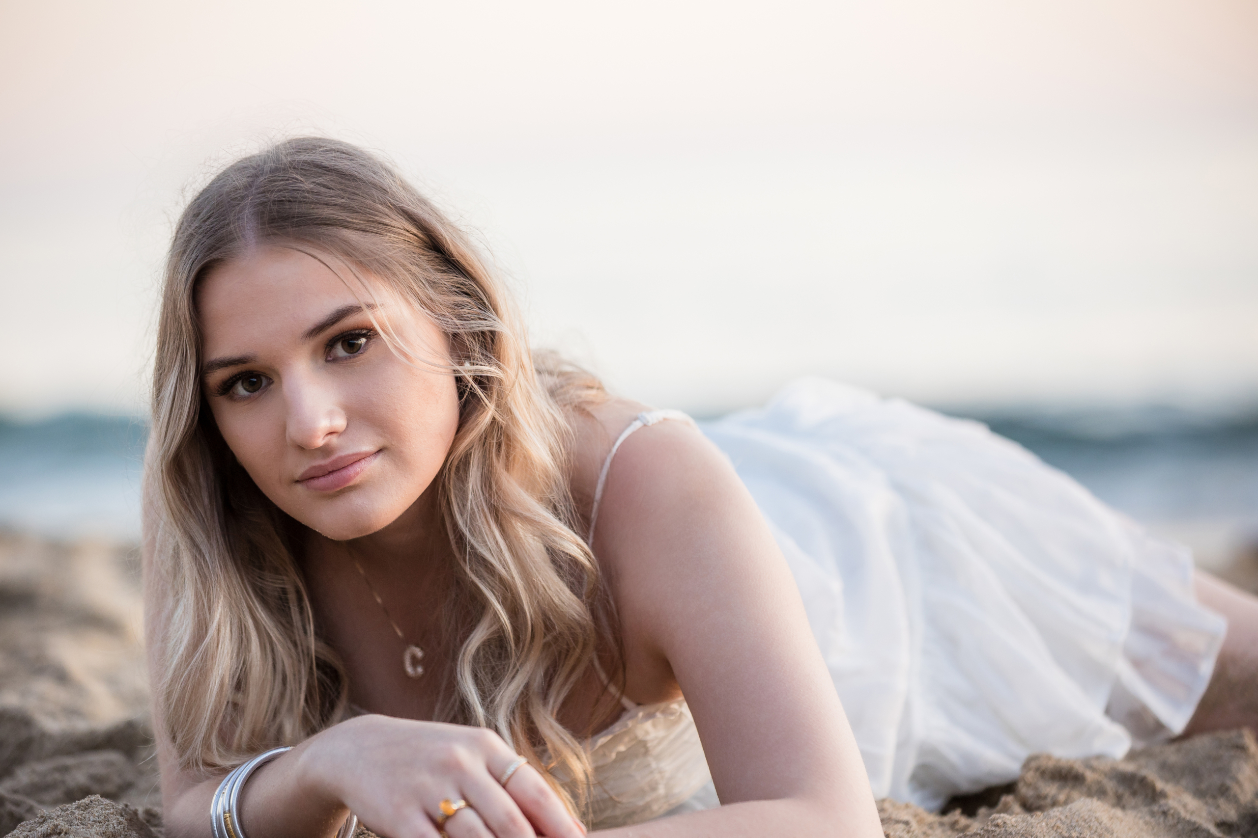 Caldwell lying on the sand in a white dress, looking directly at the camera with a serene ocean horizon behind her.
