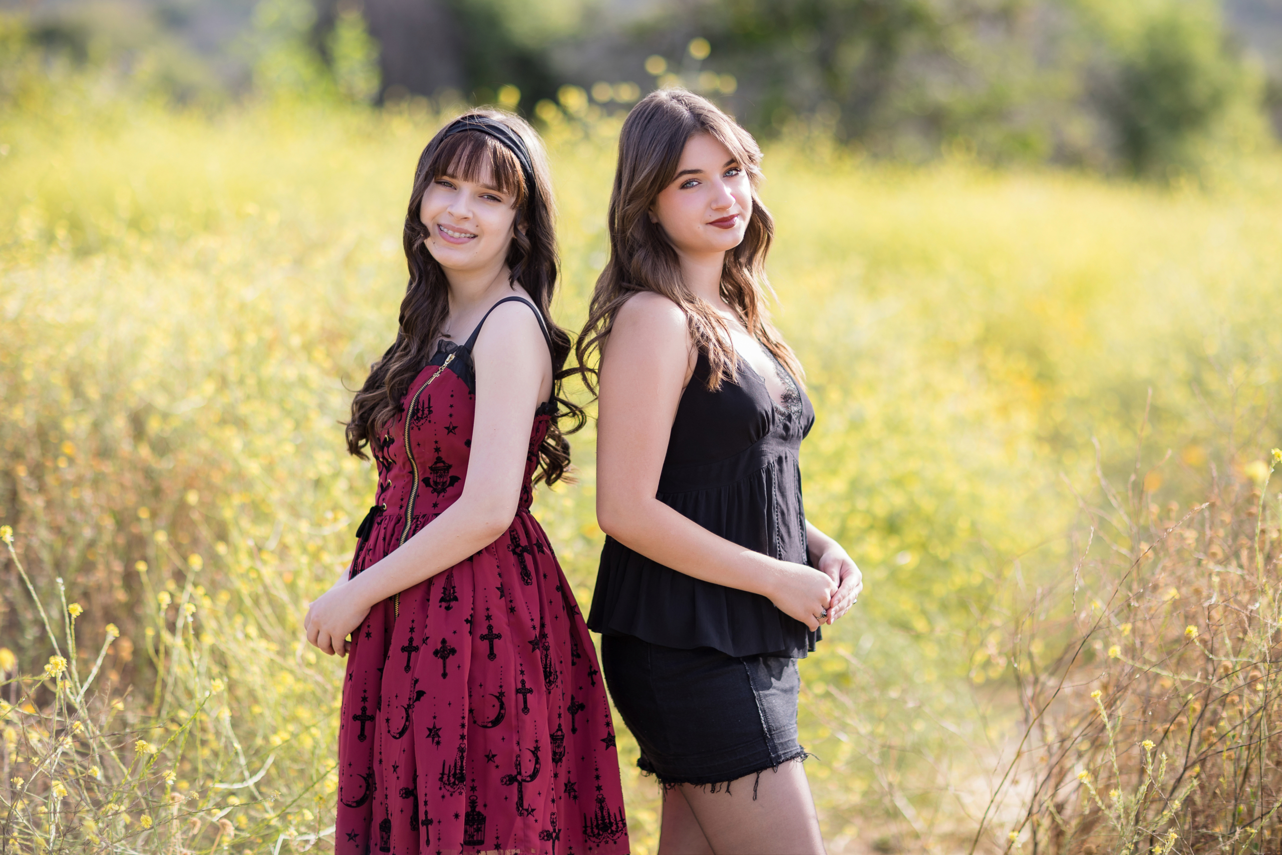 Sisters standing back-to-back in a sunlit wildflower field, one in a red dress with gothic details, the other in a black peplum top and skirt, with soft smiles and flowing hair.