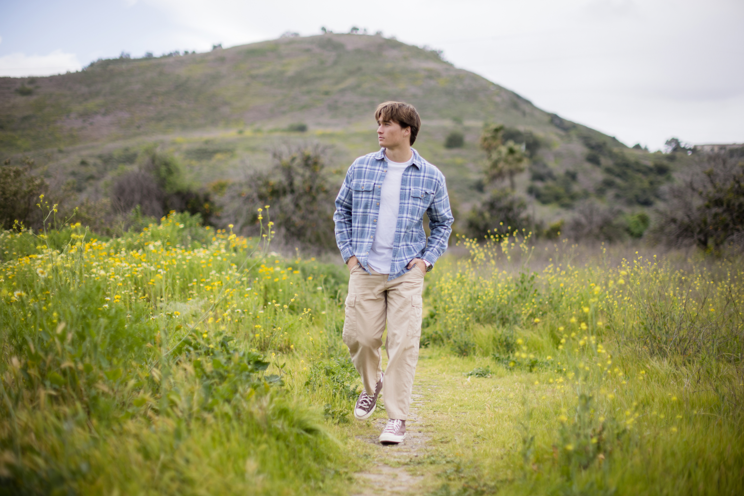 Senior guy walking along a trail through a field of spring wildflowers, with rolling green hills in the background, dressed in casual outdoor wear.