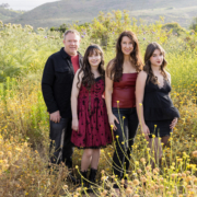 Entire family walking into a field of tall yellow wildflowers, framed by rolling green hills and springtime blooms under a bright sky.