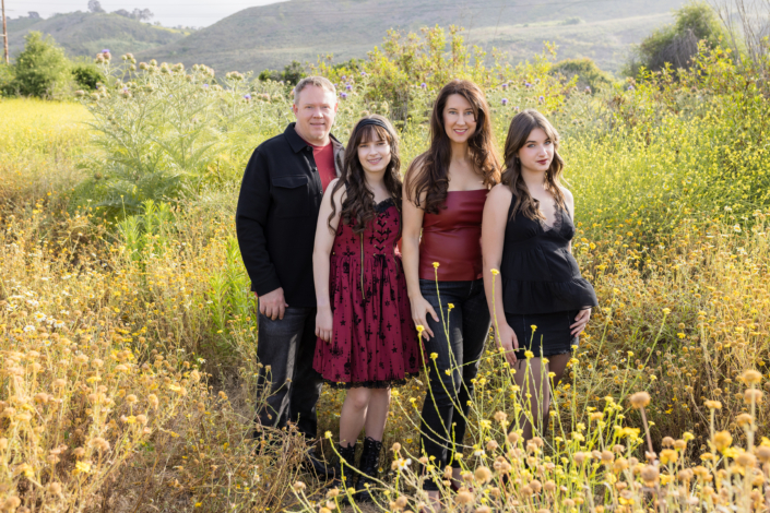 Entire family walking into a field of tall yellow wildflowers, framed by rolling green hills and springtime blooms under a bright sky.