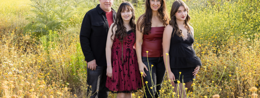 Entire family walking into a field of tall yellow wildflowers, framed by rolling green hills and springtime blooms under a bright sky.