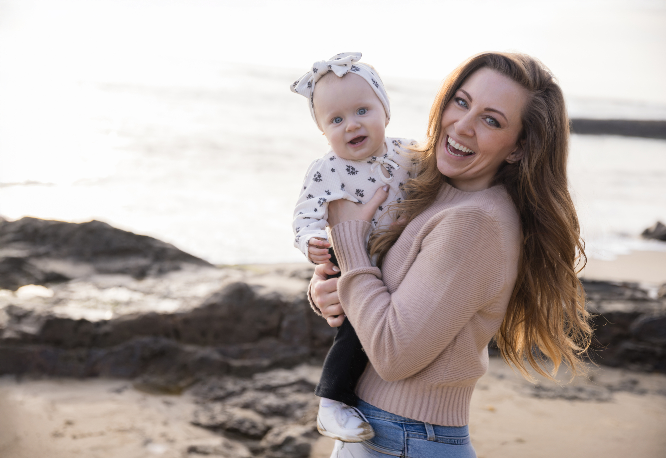 Smiling mother holding her baby daughter on a beach during a golden hour photoshoot. Both are looking at the camera with joyful expressions.