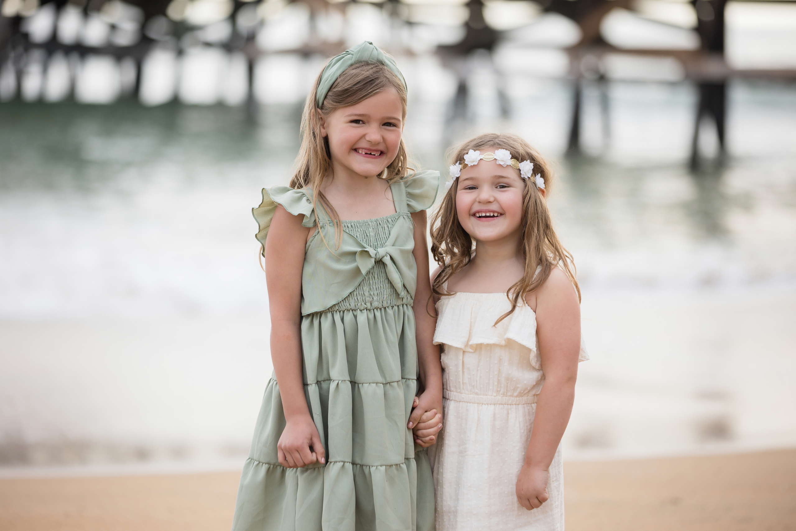 Two young girls holding hands and smiling on a sandy beach, dressed in pastel dresses with flower and headband accessories, under a pier at sunset.