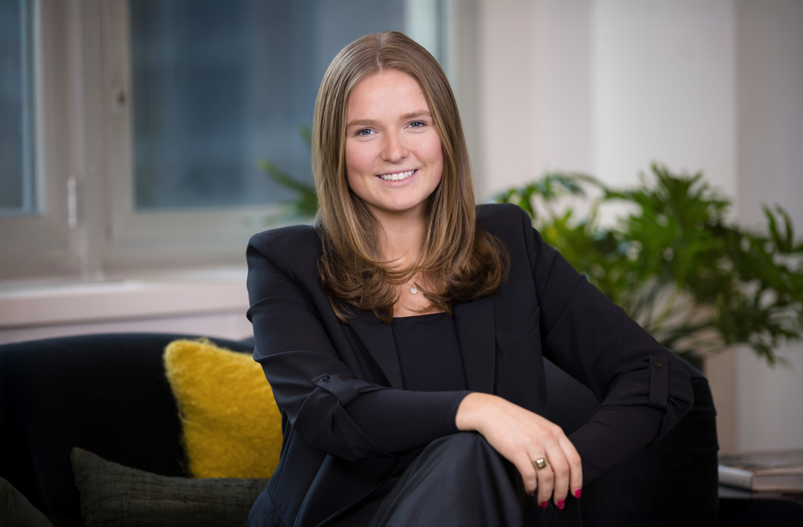 Professional woman seated in a modern office lounge, wearing a black suit and smiling at the camera