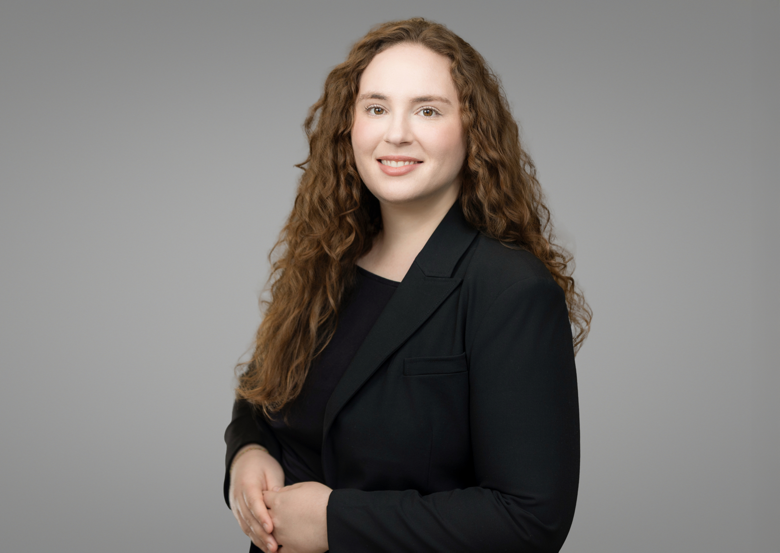 Young woman with long curly hair in a black blazer, posing against a neutral studio backdrop