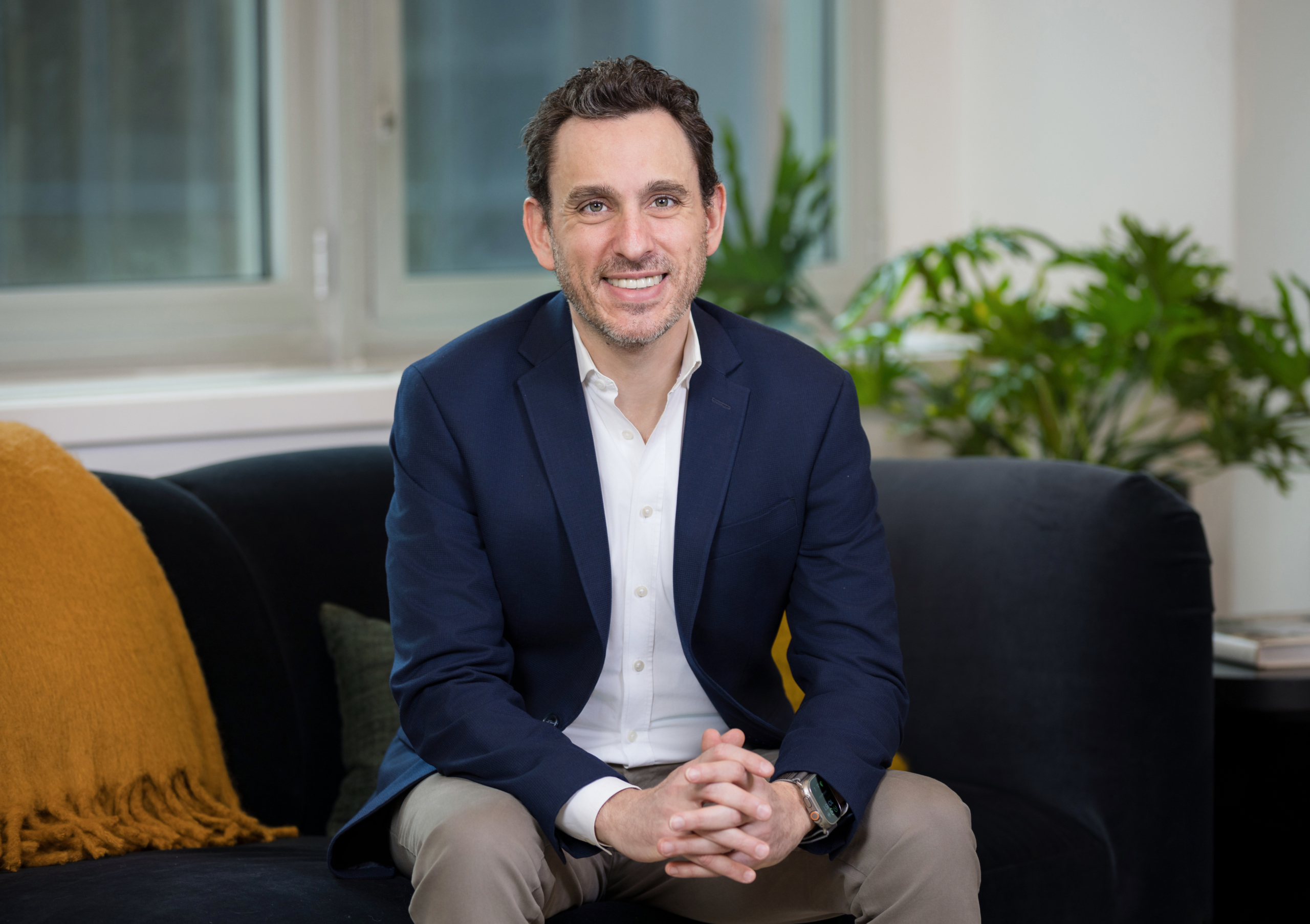 Professional man in navy blazer smiling while seated in a modern office setting with plants and natural light
