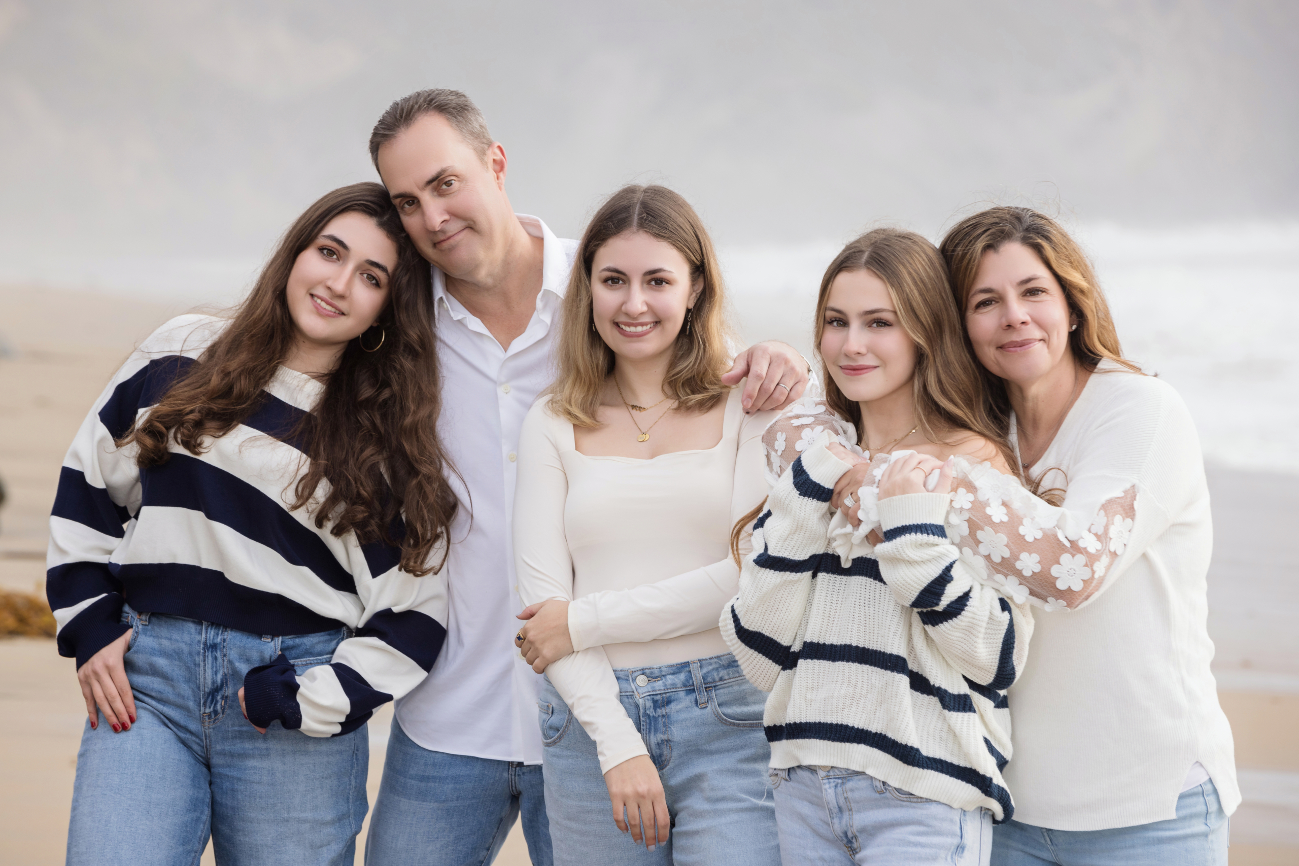 Happy family of five in casual sweaters and denim, standing arm-in-arm during a relaxed beach photoshoot near the Ritz-Carlton Laguna Niguel.