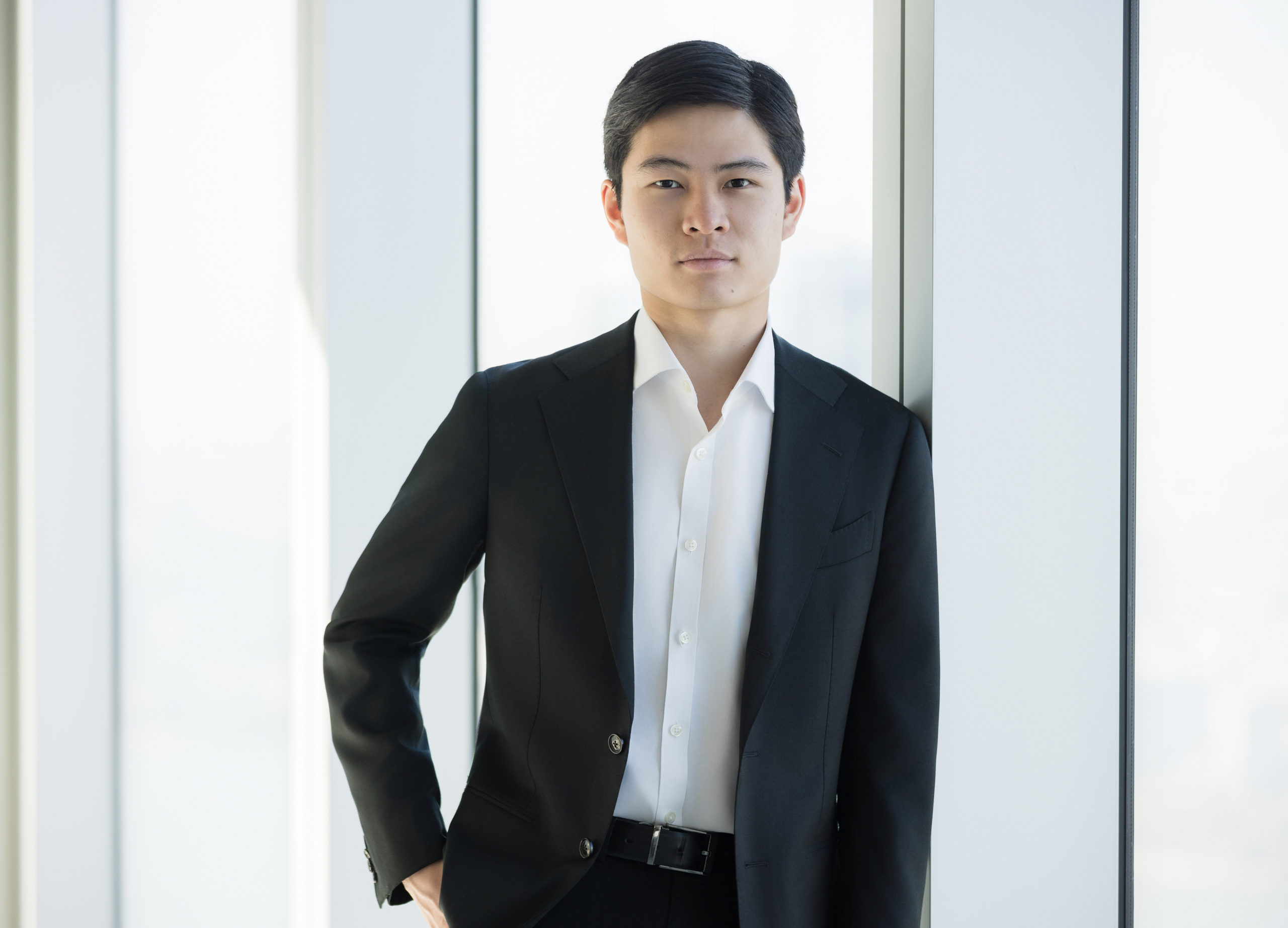 Minimalist business portrait of young Asian professional in black suit by window. client wearing classic suit from men’s headshot style guide
