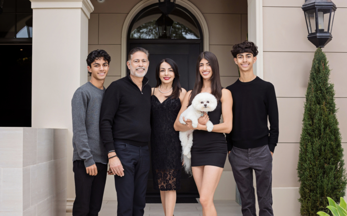 Modern family of five with their small white dog dressed in stylish black and gray attire, standing in front of their elegant home.