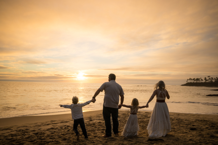 Family of four holding hands and watching the sunset during a beach photo session—capturing a warm, candid moment together.