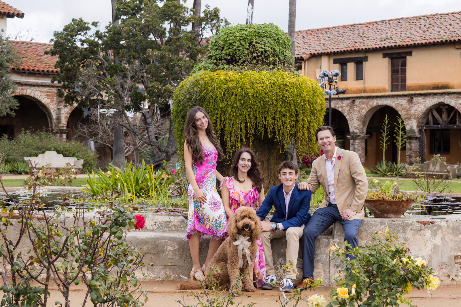 Family of four and their dog seated in the lush gardens of Mission San Juan Capistrano in summer outfits.