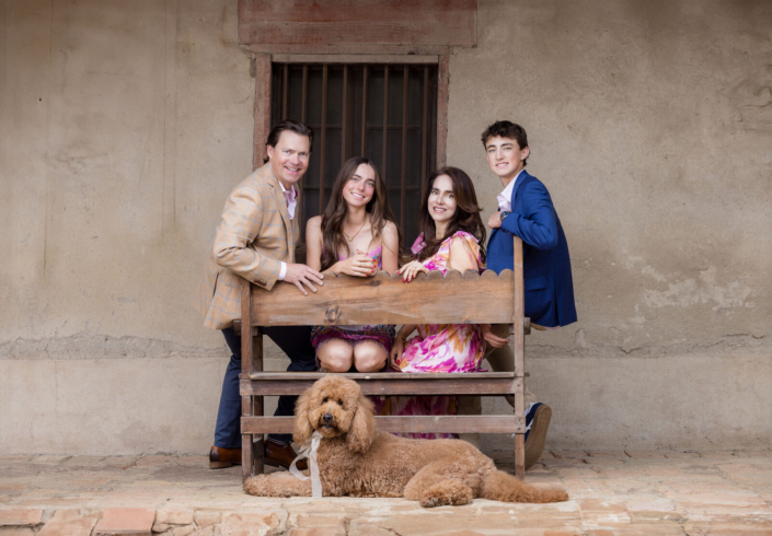 Family of four with their doodle relaxing on a wooden bench against an adobe wall at the Mission.