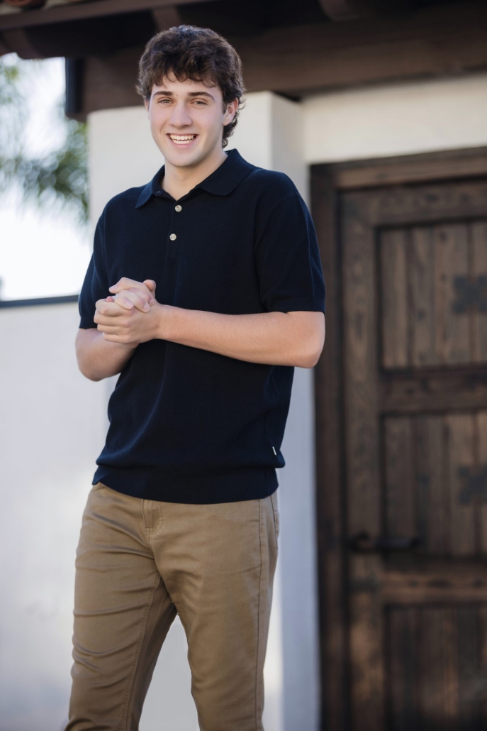 Senior guy smiling in a navy polo and tan pants under a Spanish wood-beam entry in San Clemente