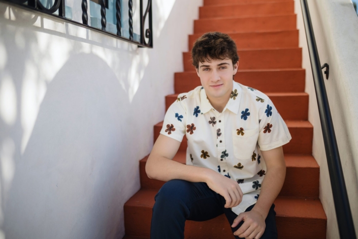 san-clemente-senior-photos-spanish-staircase-01 Senior guy in a floral shirt sitting on a terracotta Spanish staircase in San Clemente
