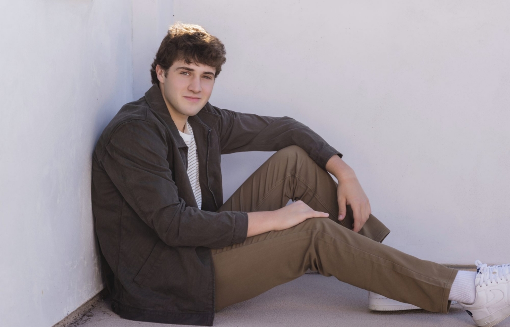 Relaxed editorial senior portrait of a guy seated against a white stucco wall in San Clemente