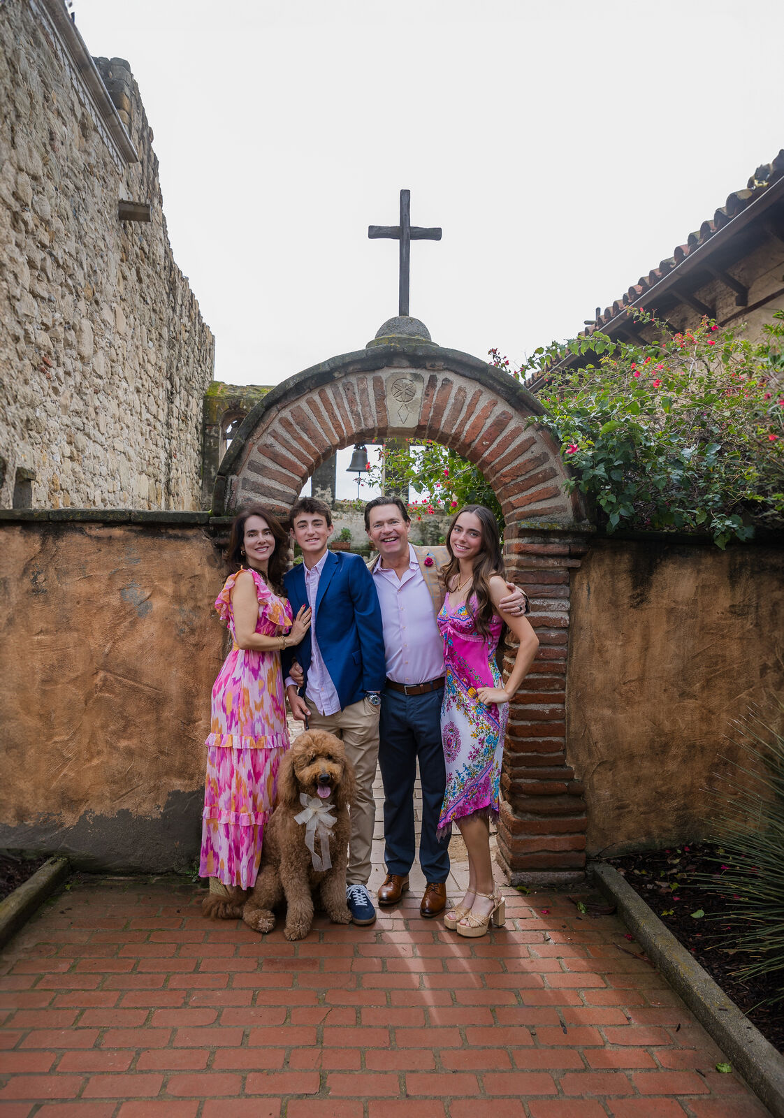 Family of four with their dog beneath the iconic cross archway at Mission San Juan Capistrano in summer florals.
