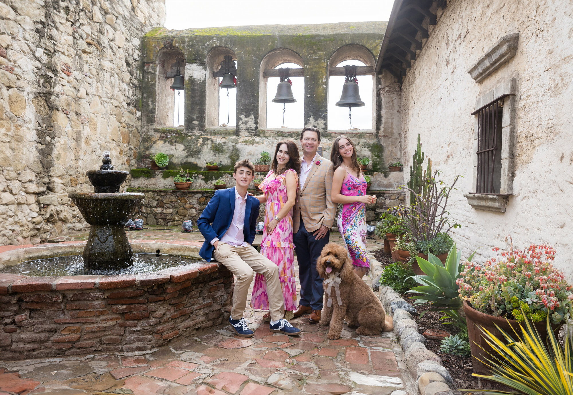 Family of four with their dog seated on courtyard steps near the Mission fountain and bells.